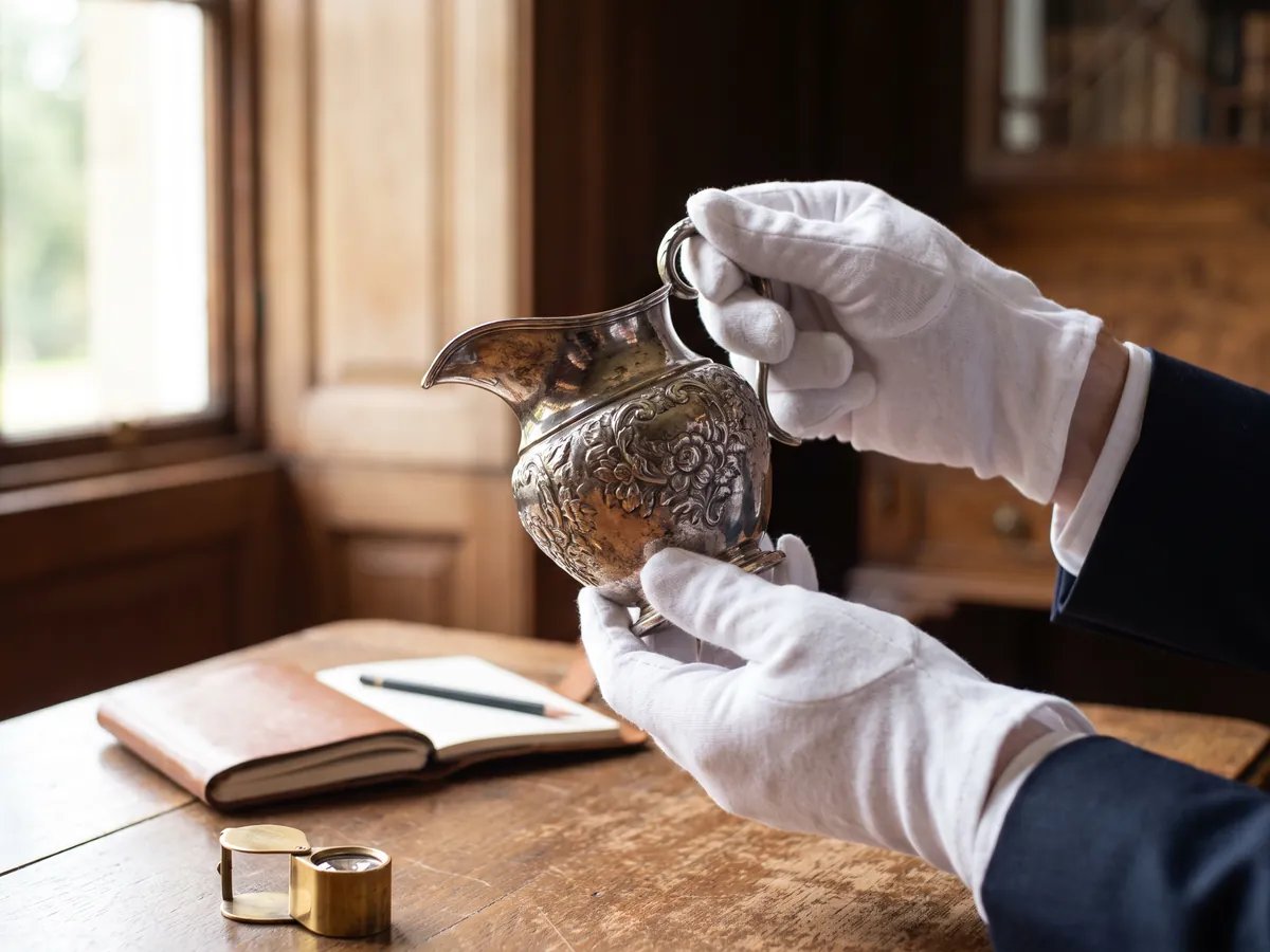 Gloved hands turning a chased silver cream jug to the light to check its condition